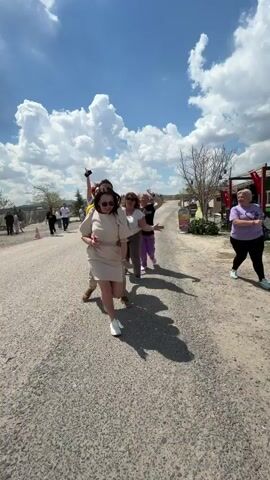 The video captures a lively group of approximately 8-10 people, primarily women, engaged in a playful follow-the-leader activity outdoors on a sunny day in Göreme, Turkey.

The scene takes place on an unpaved or lightly paved road, with a bright blue sky adorned with scattered white clouds overhead. Shadows are visible on the ground, indicating daytime, likely mid-morning or afternoon. In the background, there are sparse trees, shrubs, and what appears to be outdoor market stalls or small structures. A white tour bus is visible in the distance, suggesting a group outing.

At the beginning of the video, a man in a yellow polo shirt is at the front, playfully pushing a woman in a beige dress forward. Both are laughing and appear to be having fun. The rest of the group follows in a line, with participants smiling, waving, and making excited "woo woo" sounds. One woman, wearing a red t-shirt with the partially visible text "i m" and blue jeans, is particularly energetic, smiling broadly and gesturing as she moves. Everyone is dressed in casual attire, including t-shirts, dresses, and jeans, suitable for an outdoor activity. The overall atmosphere is one of joy, camaraderie, and lighthearted fun.