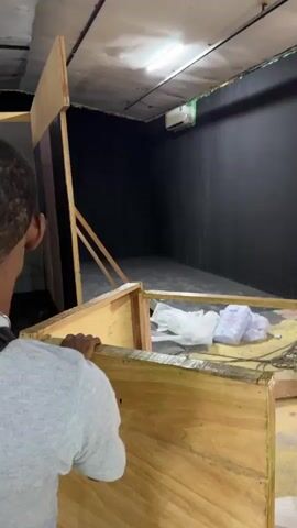 The video shows the inside of what appears to be a studio being set up. Various individuals are present, some working on scaffolding, others standing or sitting around. The studio space includes different colored walls (black, green, red and white), a mannequin, and various equipment. The atmosphere suggests preparation for a photoshoot or video shoot. The video briefly shows a person walking through, passing by a temporary wooden wall under construction. The overall environment is somewhat cluttered, with visible boxes and tools.