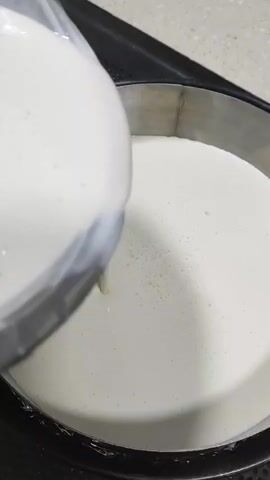 A close-up shot shows a stream of white liquid being poured from a clear glass pitcher into a round, dark-colored baking pan. The pan is filled with a smooth, off-white substance, likely batter or a similar culinary mixture, which appears to be slightly aerated. The pitcher is tilted at an angle, and the liquid flows into the pan, creating ripples on the surface. The background suggests a kitchen countertop, possibly made of a light-colored stone or laminate. The lighting is bright and even, illuminating the contents of the pan and the pouring liquid.