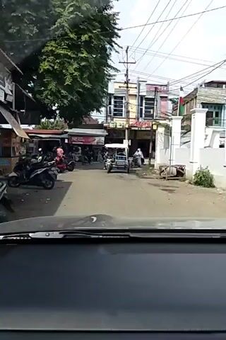 The video captures a street scene, likely in Southeast Asia, from the perspective of a vehicle's dashboard. Several motorcycles and scooters are parked along the side of the road, with one red scooter prominently featured in the foreground. Two individuals, one wearing a pink shirt and the other in a white shirt, are interacting near the red scooter. A few other people are visible in the background, some appearing to be vendors at stalls. A white car is also present on the road. The environment appears to be a bustling marketplace or commercial area, with shops and trees lining the street. The sky is bright, suggesting daytime.