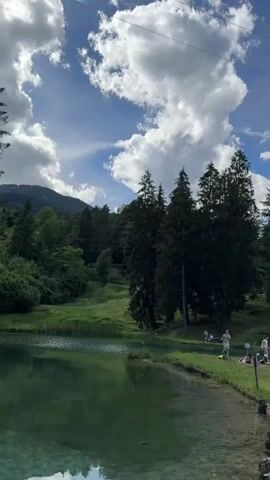 The video displays a scenic view of a lake surrounded by lush green grass and tall evergreen trees. In the foreground, the clear, turquoise water of the lake reflects the sky and surrounding landscape. People are scattered along the grassy banks, enjoying the outdoors. Some are seated, while others are standing, suggesting a relaxed atmosphere. In the background, mountains rise majestically, their peaks often shrouded in soft white clouds, adding to the picturesque setting. The sky is a vibrant blue, interspersed with fluffy white clouds that drift lazily, casting shadows on the land. The overall impression is one of a tranquil natural environment, perfect for a leisurely day out.