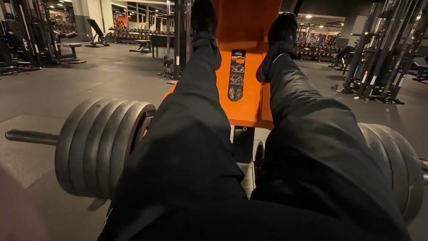 A first-person perspective video shows someone performing leg presses on a weight machine in a gym. The individual is wearing black pants and black shoes, their feet firmly planted on the orange footplate of the machine. The camera focuses on their legs and the weights, which are stacked on either side of the machine. The gym is well-equipped with various weightlifting equipment, including dumbbells, benches, and other machines, all set against a backdrop of black flooring.  The lighting in the gym is bright and even, illuminating the exercise area.