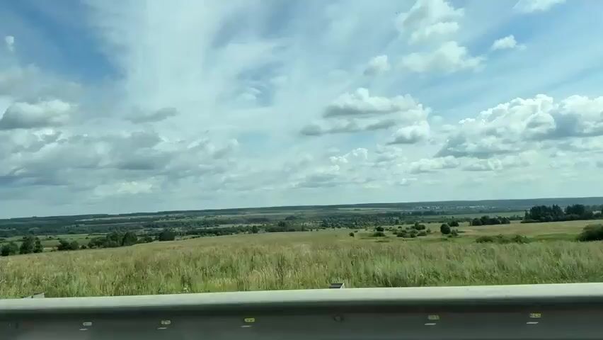 The video shows a view from a vehicle moving along a road. The foreground features a silver guardrail. Beyond the guardrail, a wide, grassy field stretches towards a distant landscape of rolling hills, trees, and scattered buildings. The sky above is partly cloudy with large, white cumulus clouds drifting across a blue expanse. The overall impression is of a rural, open environment on a clear day.