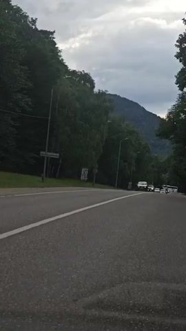 The video shows a car driving on a road through a forested mountain area. Several other cars and a bus are visible on the road ahead. The sky is overcast with some clouds. The road appears to be a two-lane highway. Trees line both sides of the road, and a mountain can be seen in the background. The time stamp indicates the video was recorded from the perspective of a moving vehicle.