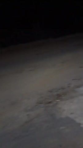 The video shows a dark, unpaved ground surface, possibly a dirt road or path, illuminated by a faint light source. The terrain appears uneven with some debris scattered on the ground. The camera is moving, giving a first-person perspective, suggesting someone is walking or driving through the area. The darkness of the surroundings and the limited visibility indicate it is nighttime.