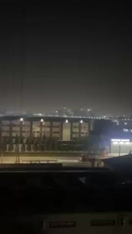A nighttime view shows an urban or industrial area with several buildings. In the foreground are the rooftops of lower structures, and in the background is a larger, illuminated building with multiple windows and a row of trees along its base. Lights from other buildings and possibly distant city structures are visible in the hazy background. A truck drives along a road in front of the large building. Power lines stretch across the frame in the upper left quadrant. The overall scene is dimly lit, creating a sense of quiet and stillness.