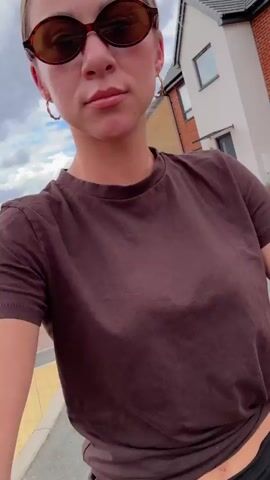 A medium shot captures a woman with blonde hair pulled back, wearing oval brown sunglasses, gold hoop earrings, a brown t-shirt, and a black skirt. She is outdoors on a paved path, possibly a sidewalk, with a street in front of her and houses in the background. The sky is overcast. The woman's head and upper body are visible, and she appears to be looking at the camera. The video is shaky, suggesting it might have been filmed while walking or riding.