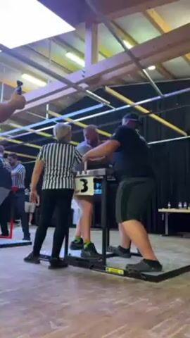 The video shows two men arm wrestling on a stand with a referee standing close by. The man on the left has a gray beard and is wearing a light gray t-shirt, the other man is wearing a black t-shirt and gray shorts. The referee is wearing a black and white striped shirt. The arm wrestling stand is labeled "A.A.F." in white lettering on a black background. A small table is set up behind them with water bottles. There are a few other men in the background, and someone is filming the arm wrestling with their phone. The environment appears to be an indoor event space.