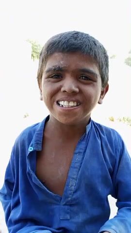 A young boy with dark skin, short black hair, and a blue collared shirt is looking directly at the camera. His mouth is open and he appears to be speaking or laughing. The background is out of focus and appears to be outdoors with some vegetation.