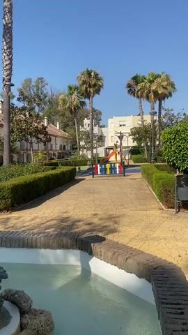 The video opens on a view of a park-like setting with a fountain in the foreground, its water feature active. Behind the fountain is a paved walkway, lined with manicured hedges, and further back, a playground with colorful equipment. In the distance, rows of multi-story buildings with balconies and windows are visible, interspersed with palm trees and other foliage. The sky is clear and blue, indicating a sunny day. The camera pans slightly to focus on the fountain's statue, which depicts a figure pouring water from an outstretched hand.