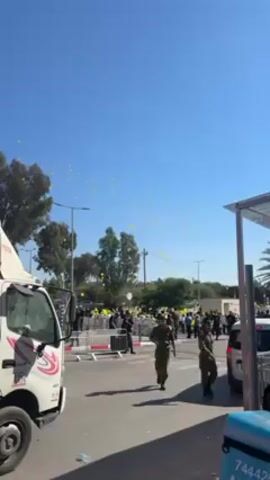 Here’s a description of the video:

The video shows a crowd of people gathered on a sunny day, with many yellow balloons floating in the sky. A white truck with "TOYOTA" written on it is parked on the left side of the frame. Several people wearing dark clothing appear to be maintaining order in the crowd. One man in military-style clothing walks purposefully from the background towards the foreground of the shot. There is some sort of metal barrier separating the crowd from the street.  The environment includes trees, street lights, and a partially visible building structure on the right side of the frame.