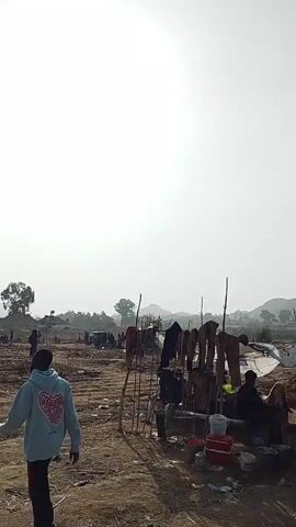 The scene is an outdoor area that appears to be a rural or underdeveloped location with a bright, overcast sky. The ground is dry and dusty, with sparse vegetation. In the foreground, there's a green and yellow auto-rickshaw, a small makeshift shelter or stand with clothes hanging on poles, and some buckets and containers.  

Further into the scene, there are numerous people scattered across the landscape.  They appear to be engaged in some kind of work, possibly digging or sorting through earth. A motorcycle and a dark-colored car are parked nearby. In the background, there are rolling hills or small mountains. The overall impression is of a busy, perhaps impoverished, work site.