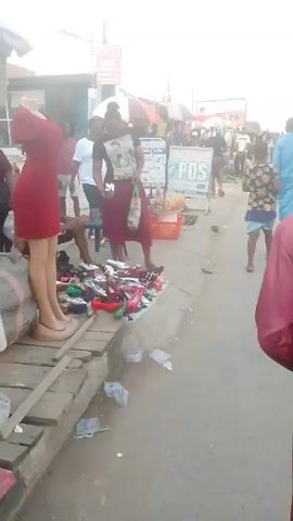 The video shows an outdoor street scene, likely a market or commercial area. The initial shot focuses on a display of various women's shoes spread out on the ground, near a mannequin wearing a red dress. A woman wearing a maroon skirt and t-shirt is standing near the shoe display, possibly examining them.  Several people are visible in the background. The camera then moves, following a young man wearing a red shirt. The perspective shifts to show the street, which is bordered by shops and vendors under umbrellas. People are walking along the sidewalk, and there are cars and a yellow three-wheeled vehicle on the road. The general atmosphere is busy and active, indicating a lively commercial area.