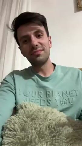 The video shows a medium close-up of a man with dark hair, wearing a light green sweater with the words "YOUR PLANET NEEDS YOU" emblazoned on the front. He is sitting down, holding a fluffy, olive-green pillow in his lap. The background includes a white curtain and a framed picture hanging on the wall. The man rubs his face and nose with his hands, then speaks. The lighting is soft and natural.