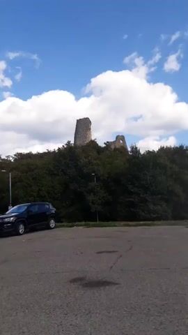 The video begins by showing a distant view of ruins on a hilltop against a backdrop of blue sky and white clouds. In the foreground, two dark-colored SUVs are parked on a gravel surface. A blue signpost is visible to the left of the vehicles. The camera then pans to the right, revealing more of the landscape, including trees and more signs. An orange vehicle is parked further down the road. As the video continues, the camera moves, showing a closer view of the road and trees lining the side. The ruins on the hilltop remain visible throughout the short clip. The environment appears to be an outdoor setting with a road, vehicles, and natural elements.