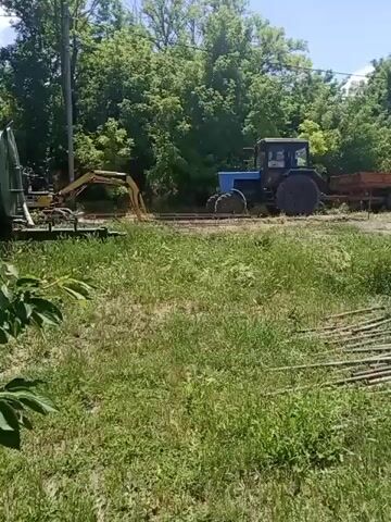 A blue tractor with a trailer is parked on a dirt path, next to a yellow excavator. Tall green trees line the background, and a clear blue sky is visible above. In the foreground, tall green grass and a pile of wooden poles are scattered across the ground. The video is shot outdoors in daylight.