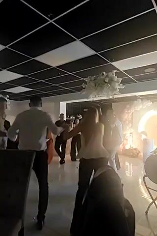 The video shows a group of people dancing in a room with a black and white checkered ceiling. White floral decorations hang from the ceiling and a large, illuminated circular archway is visible in the background. The lighting is warm and festive. People are seen holding hands and dancing in a circle or in pairs. The attire suggests a celebratory event.