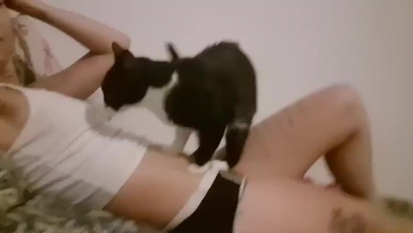 A black and white cat is positioned on the stomach of a person lying down. The person is wearing a white tank top and dark underwear. The cat appears to be kneading or gently pawing at the person's stomach. The person's arm is raised and resting behind their head. The background suggests a bedroom setting with pillows and a patterned blanket.