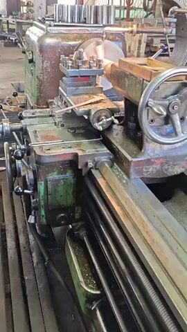 A metal lathe is shown in operation in a workshop environment. A rotating workpiece is being machined by a cutting tool, with coolant being applied to the cutting area. Metal shavings are visible accumulating around the workpiece and the lathe. The lathe itself appears to be older, with signs of wear and tear. The background suggests a cluttered workshop with various tools and materials.
