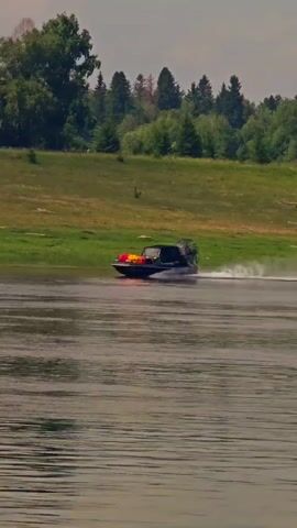 A hovercraft speeds across a wide body of water, kicking up a spray as it moves. The hovercraft is dark colored with yellow and red accents on its bow. In the background, a grassy hill slopes upwards, dotted with trees. The sky is overcast.