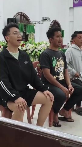 The video shows a group of men sitting on chairs in what appears to be a church or a hall. They are all facing forward and singing or speaking with their mouths open. In the background, there is a wooden altar decorated with white flowers and green fabric. A keyboard is visible in the foreground, with someone's hands on the keys, suggesting they are accompanying the singers. The men are dressed casually in t-shirts and shorts or pants. The lighting is bright, illuminating the room. The overall atmosphere seems to be a communal gathering or a religious service.