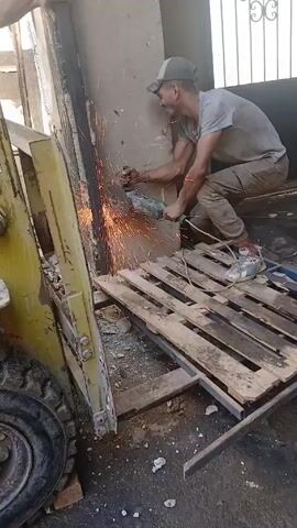 A man in a grey t-shirt and tan pants is using a handheld angle grinder to cut metal attached to a concrete wall. He is wearing a cap and sneakers. Sparks are flying from the point of contact between the grinder and the metal. He is crouching on a wooden pallet that is resting on a metal frame. Next to him is a yellow machine with a tire. An arm appears in the top left corner of the frame, possibly assisting in stabilizing the metal being cut. The setting appears to be an outdoor work area or construction site. The wall has a dark vertical streak on it. Part of a gated window is visible in the background.