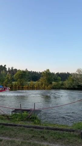 A red boat with a blue canopy and several people aboard is moving quickly across a body of water. The boat is kicking up a white spray as it moves, creating ripples and waves in the water. The background shows a dense line of green trees and foliage under a clear blue sky. In the foreground, there is a grassy bank with some wooden structures and red rope fencing.