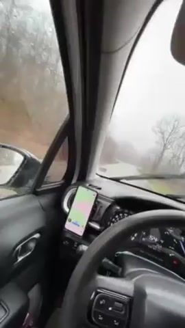 A first-person view from inside a car shows a drive on a wet and foggy day. The windshield wipers are on, and a phone mounted on the dashboard displays a map. The car is driving on a winding road through a wooded area with bare trees. The car's speedometer is visible, showing speeds fluctuating between 52 and 56 km/h. Music is playing in the background.