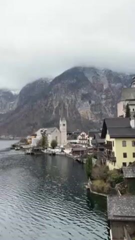 The video shows a picturesque lakeside town nestled against a backdrop of tall mountains capped with a dusting of snow. The sky is overcast and gray, creating a soft, diffused light over the scene. The calm, dark water of the lake reflects the town and mountains, adding to the serene atmosphere. The town itself is made up of buildings with traditional architecture, including a prominent church with a tall steeple. There's a boat in the water, and the houses are on the waterfront.