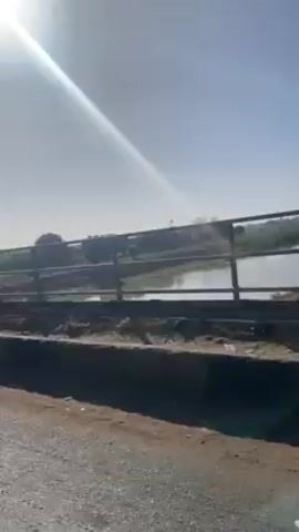 The video shows a view from a vehicle crossing a bridge. The landscape features a body of water, possibly a river or a wide stream, with vegetation and patches of land interspersed within it. The bridge has a simple metal railing. The weather appears sunny and clear. The road surface is visible in the foreground. The vehicle is in motion, as indicated by the changing perspective. There are no people clearly visible in the video. The focus is on the environment and the crossing of the bridge.