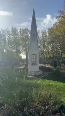 The video shows a narrow, white, triangular structure with a pointed roof standing in a grassy area. There are two visible windows on the structure, one higher up and one lower down. The structure is surrounded by trees with leaves that suggest it is autumn. The background shows a street with cars and buildings. The day appears sunny with a few clouds. The camera is static, providing a clear view of the structure.