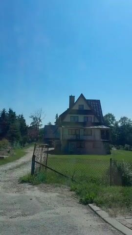 The video shows a dirt road leading towards a cluster of houses nestled among trees. The sky is clear and blue. The road curves to the right. A fence with a gate is visible on the right side of the road. One house, which appears to be made of wood and stone, is prominently featured. It has a steep, gabled roof and multiple windows. Other houses and trees are visible in the background.