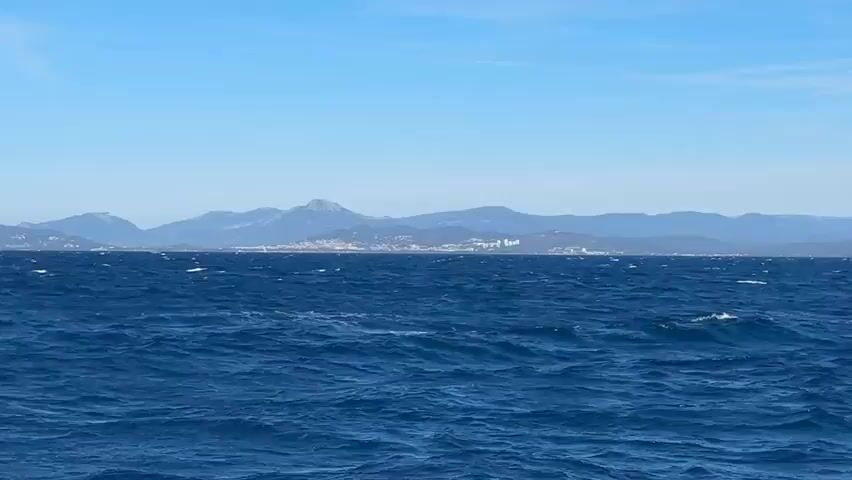 The video shows a wide shot of the ocean under a clear blue sky. The water is a deep blue and appears choppy, with whitecaps indicating a moderate wind. In the distance, a coastline is visible with rolling hills and mountains. A town or city is built along the coast, with buildings dotting the landscape. The sun appears to be high in the sky, casting bright light on the scene. The camera is stationary, offering a static view of the seascape. There are no people or vessels visible in the frame. The overall impression is one of a vast and open ocean meeting a distant, mountainous landmass.