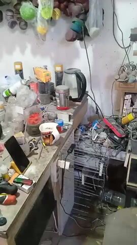 The video pans across a cluttered garage or workshop. The scene is dimly lit, with a mix of natural and artificial light sources. In the foreground, a workbench is covered with various items, including tools, bottles, jars, and a mobile phone. A white mug sits on the left side of the workbench. Further back, a white refrigerator stands against the wall. To the left of the refrigerator, car seats are stacked. In the middle of the scene, a blue hatchback car is parked with its trunk and driver's side door open. Various tools and equipment, including a blue fan and coiled wires, are scattered around the car and on the floor. The overall impression is one of a disorganized but functional workspace.