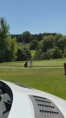 A golf course is shown on a sunny day. The video is shot from inside a golf cart, as evidenced by the vehicle dashboard visible at the bottom of the frame. A person is walking away from the camera, pushing a golf bag on a wheeled cart along a path on the golf course. The person is wearing a light blue shirt and a light-colored skirt with a floral pattern. The golf bag is black. The golf course is green, with trees and bushes visible in the background. The sky is blue.