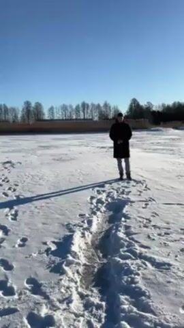 A man is walking on a frozen, snow-covered lake on a sunny day. He is wearing a black coat, blue jeans, and tan boots. He slips on some ice and stumbles, but manages to regain his balance.  There are trees visible in the background, along the edge of the lake. The sky is clear and blue.  Footprints are visible in the snow.  There is a dark, shallow track of slush and water where the ice has partially melted.