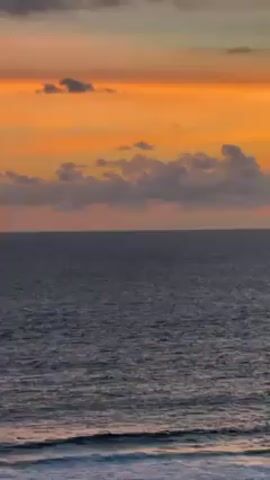 The video shows a view of the ocean at sunset. The sky is a mix of orange, yellow, and grey clouds. The ocean is a dark blue, with small waves visible near the shore. A small boat is seen in the distance.