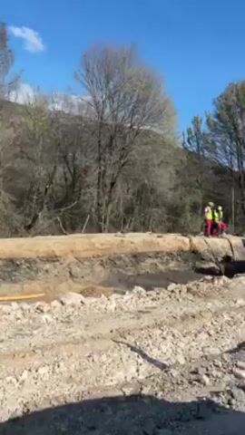 The video shows a construction site on a sunny day. In the background, there's a line of trees with a few bare branches. In the foreground, there is a construction pit with a yellow pipe visible. An excavator is filling a dump truck with soil from the pit. There are two construction workers wearing yellow vests and red jackets standing near the pit. A red and white striped barrier is visible in the foreground.