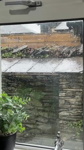 The video shows a view through a closed window on a rainy day. Outside, a stone structure with a slate roof is visible, covered in raindrops. Some greenery is growing along the edges of the roof. The window is framed in white, and a small potted plant sits on the windowsill to the left. Raindrops are visible on the windowpane, obscuring the view slightly.