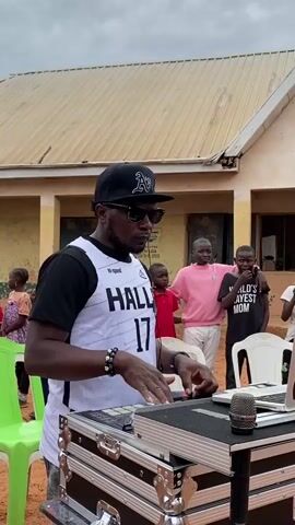 A man in sunglasses and a baseball cap works on a laptop placed atop a black DJ console, a microphone in front of it. He wears a white jersey with black sleeves and a black and white design. Behind him stand several children, one in a pink shirt and another in a black shirt that reads "WORLD'S KAYEST MOM." The backdrop is a building with a sign stating "2013 MDGs CGS-JOS-SOL LEA PRIMARY SCHOOL DAKYEN KURU JOS." Some green plastic chairs are also visible. The environment appears to be outdoors.