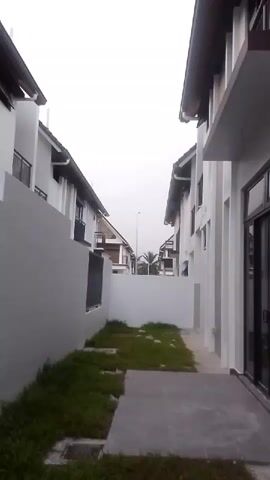 The video shows a side view of a narrow walkway between two modern houses. The houses are white with brown trim and appear to be two stories high. There is a patch of grass on the left side of the walkway, with a drainage grate visible. The sky is overcast. The video is stationary.