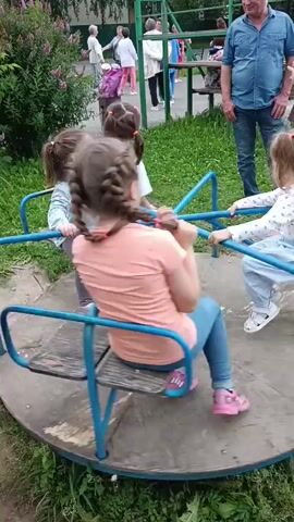 The video opens on a merry-go-round with at least four children on it. The merry-go-round is blue and gray, with wooden seats. The children are all young girls, and they are wearing casual clothing. The girl in the foreground is wearing a white t-shirt with a flower on it and pink shorts. She has her hair in two pigtails. The girl to her left is wearing a white t-shirt and gray leggings. She also has her hair in pigtails. The girl to her right is wearing a pink t-shirt and has her hair in two braids. The fourth girl is partially visible at the back of the merry-go-round. In the background, there are adults standing around. There is also some greenery, suggesting the merry-go-round is in a park or playground. The camera pans slightly, showing more of the background and the other children on the merry-go-round. The scene appears to be a sunny day.