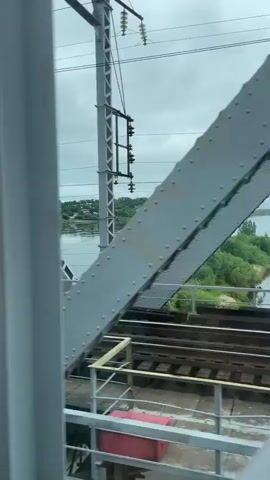 The video shows a view from a moving train crossing a bridge over a river. The bridge is a steel truss bridge. The river is wide and calm, with green trees and some buildings on the far bank. The sky is cloudy and overcast. The train tracks are visible in the foreground. There is a small island in the river with trees. A red object, possibly a trash can or marker, is visible on the embankment near the bridge.