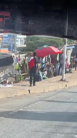 The video, shot from inside a car, shows a bustling street scene. People are walking along a sidewalk lined with small stalls selling various goods. Some of the stalls are covered with red and green umbrellas. The camera pans to show more of the street, revealing a motorbike with a driver wearing a green vest, laden with large packages. The overall environment is crowded and active.