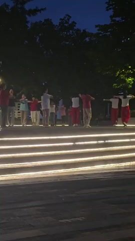 A group of people are performing synchronized movements in an outdoor setting at dusk. They are arranged in a line, facing away from the camera, with some individuals dressed in red pants. The background features dark trees and a dimly lit sky. In the foreground, illuminated by warm lights, are horizontal steps. The individuals appear to be engaged in a form of exercise or dance.