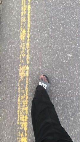 A first-person perspective shows someone walking on a paved road. The person is wearing black pants and grey KARL Lagerfeld branded slide sandals. A yellow line runs along the left side of the road. As the view pans up, a street and buildings can be seen in the distance. There's green grass and a few trees to the left of the road and streetlights along the roadside. A figure on a motorcycle is far ahead on the road. The sky is overcast.