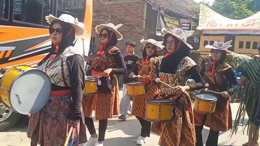 A group of young people are marching in a parade. The first few individuals are girls wearing patterned tops, red headscarves, straw hats with decorative elements, and elaborate skirts. They are each playing a small drum with drumsticks. Behind them are boys wearing costumes made of green palm leaves, resembling a tall headdress and a skirt-like garment. Two of these boys are carrying a long wooden pole with red ends on their shoulders, with other individuals also in similar leaf costumes. In the background, there is a large orange and white bus, a building with a sign, and what appears to be a residential area. The scene is outdoors on a sunny day.
