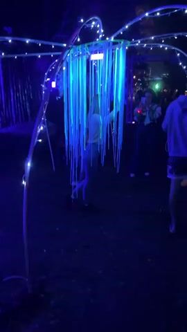 The video shows a nighttime outdoor scene illuminated by blue and white lights. In the center, a person is partially obscured by hanging blue and green streamers, positioned within a circular frame adorned with white string lights. The structure resembles a large, whimsical art installation, possibly part of a festival or event. Several other people are visible in the background, also interacting with the illuminated environment. The overall atmosphere is festive and slightly mysterious due to the low light and vibrant colors.
