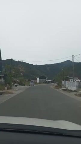 A car's dashboard is visible in the foreground of the shot, suggesting the perspective is from within a moving vehicle. The vehicle is driving on a paved road, which curves slightly to the left. On the left side of the road, there are buildings and a flag with red and white stripes, appearing to be the Indonesian flag, is mounted on a pole. A few vehicles, including a silver car and a motorcycle, are parked or moving on the road. The right side of the road has a white fence and some vegetation. In the background, there are rolling hills covered with trees, and the sky is overcast. The overall impression is a rural or semi-rural setting with some signs of human activity and infrastructure.