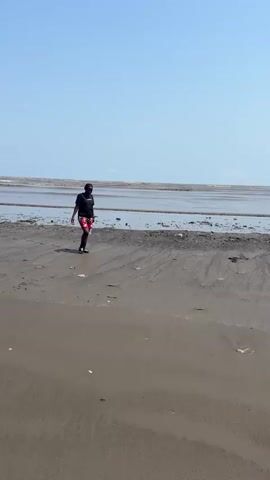 A woman walks towards the camera on a sandy and muddy beach or coastline, holding a clear plastic water bottle. She wears a black t-shirt with a "WONDER" logo and what appears to be "ENGINEERING" or "SOLUTIONS" written below it, patterned shorts (red, black, and white), black socks, and black sneakers. The sky is clear and bright blue. As she nears the camera, she stops, smiles warmly, and looks directly at the viewer. In the background, there's a wide expanse of shallow water or mudflats, with faint waves visible further out. Some debris is scattered across the ground. The scene is set during the daytime in Douala VI, Cameroon.